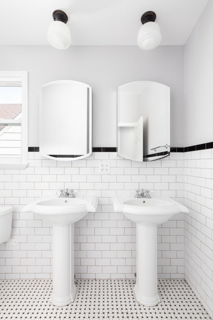 A bathroom with two pedestal sinks, tiled floor and wall, and lights hanging above.