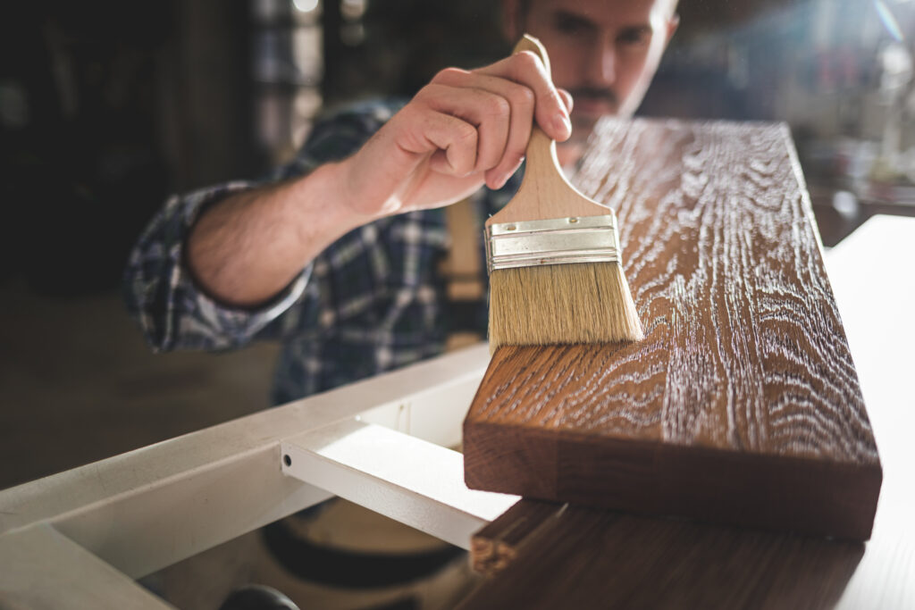 Close up of paintbrush applies paint or varnish on wooden board in carpentry workshop