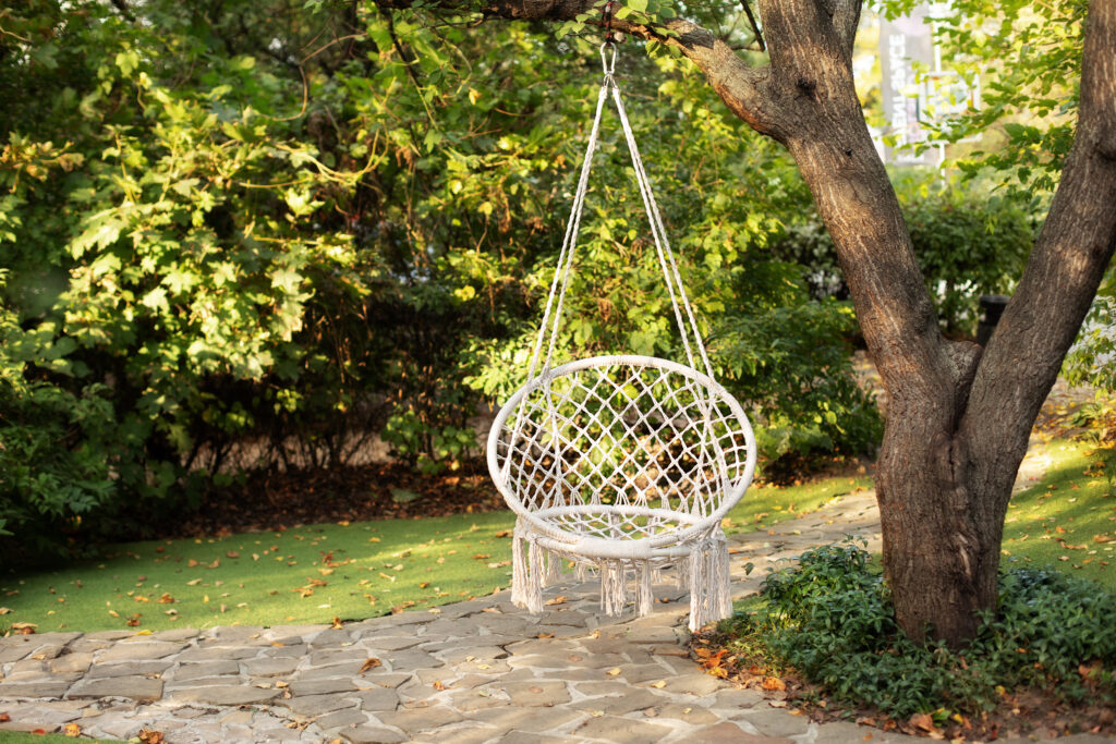 Comfortable hanging wicker white chair in summer garden. Cozy hygge place for weekend relax in garden. Hammock chair in boho style hanging on tree. Cozy exterior backyard. Concept of rest outdoor.