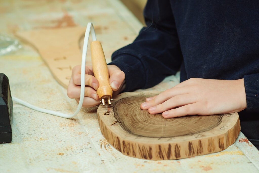 Kid makes wooden clock in the workshop. Boy burn out numbers with soldering iron on wooden disc.