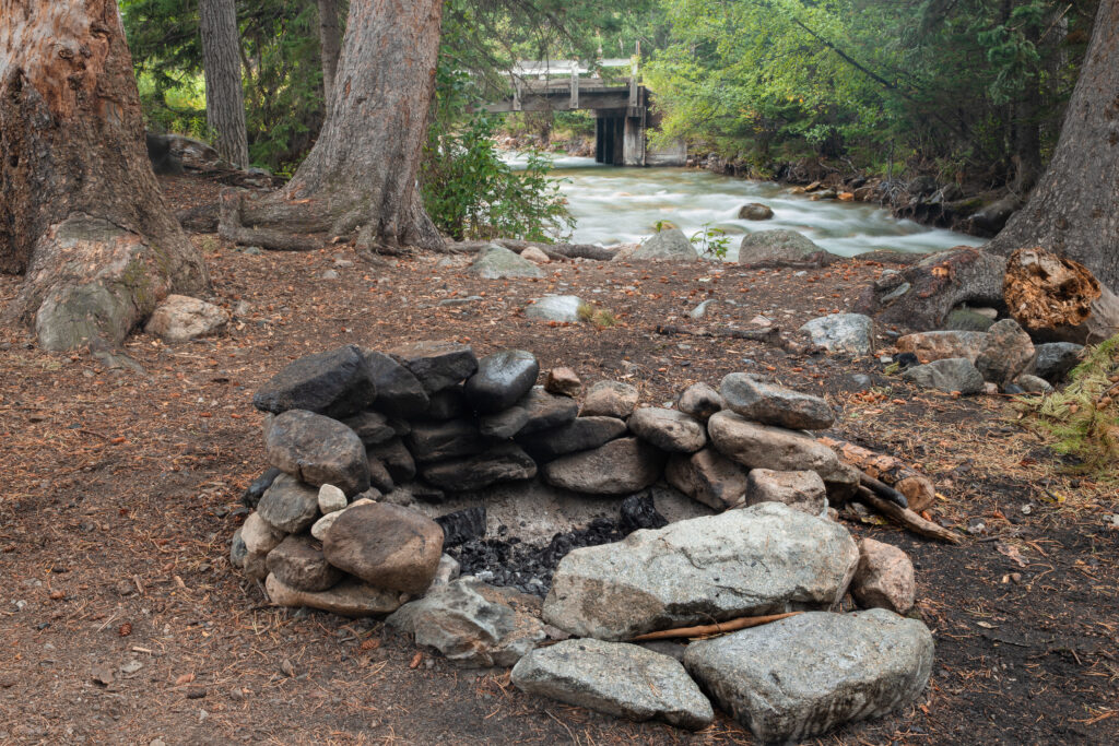 Stacked rock wall fire pit at makeshift camp site in forest woods to use as campground for camping with safety to prevent forest fire in tree clearing near river and bridge