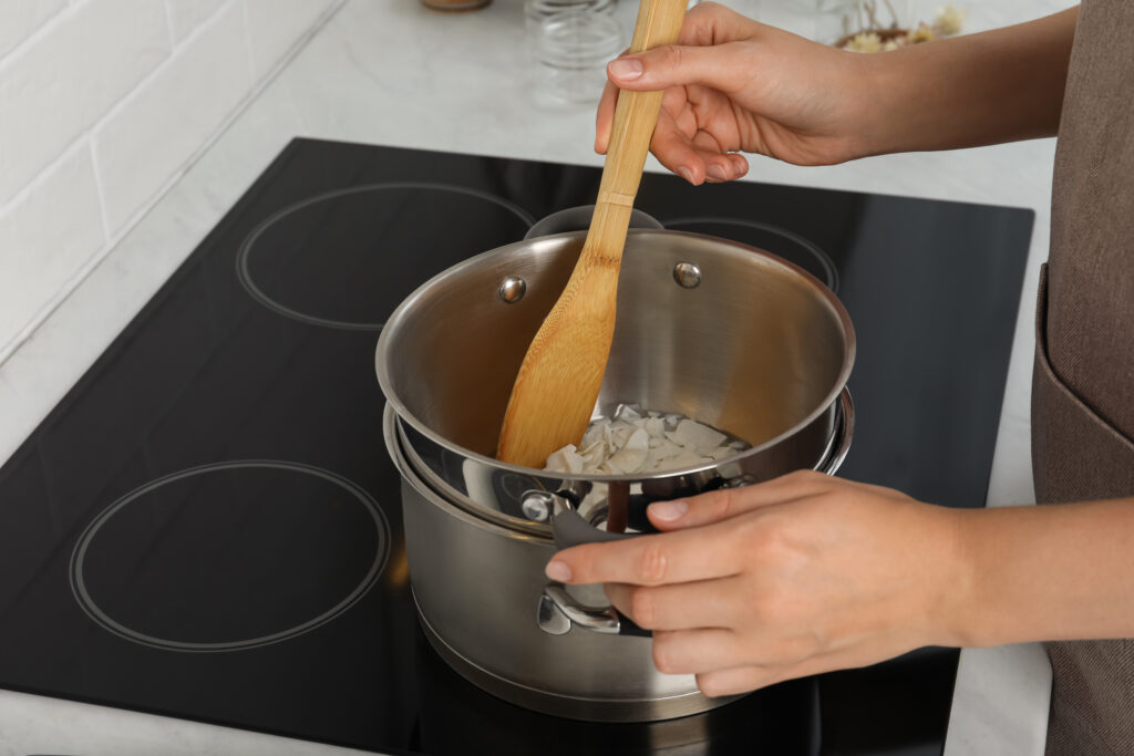 Woman melting wax on stove in kitchen, closeup. Making homemade candles