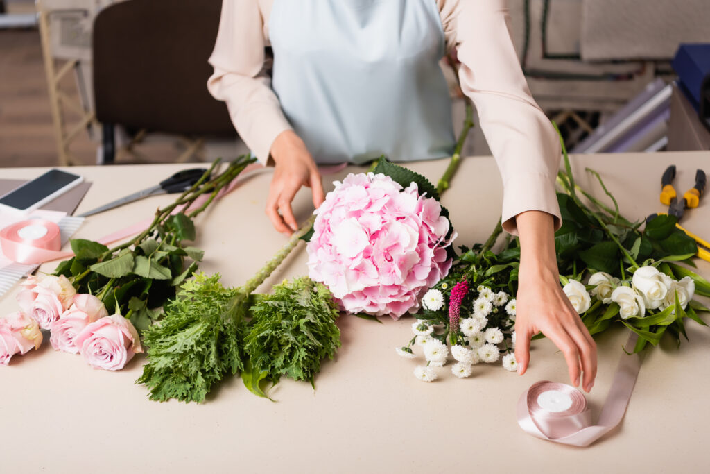 Cropped view of florist taking decorative ribbon near flowers on desk with tools on blurred background