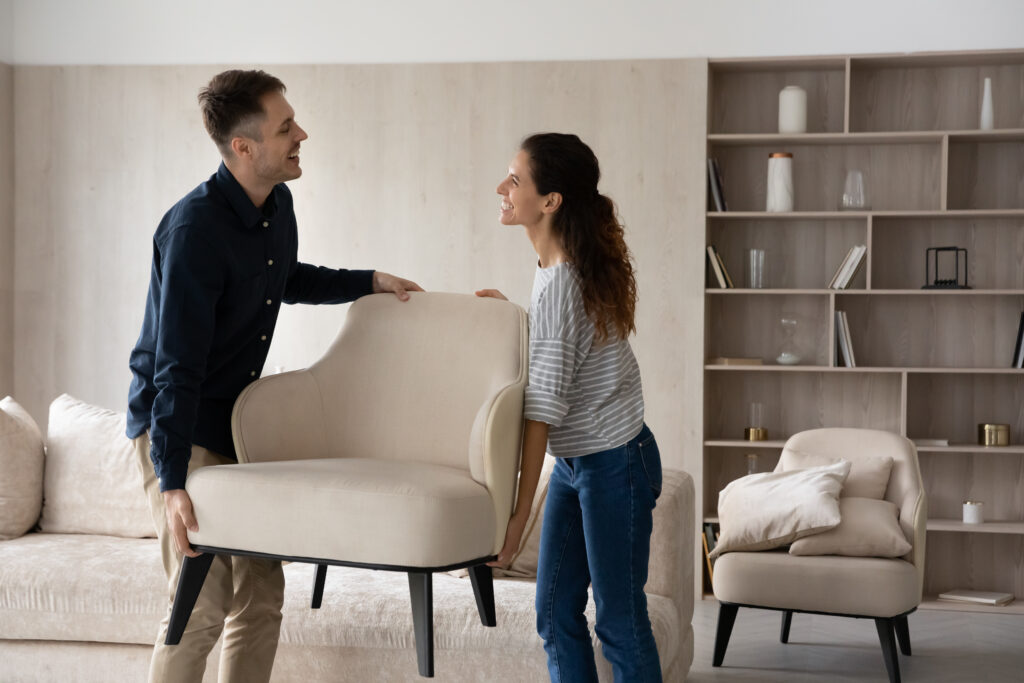 Happy beautiful hispanic young family couple homeowners holding armchair, enjoying renovating living room rearranging furniture together. Smiling spouses relocating on moving day into own dwelling.