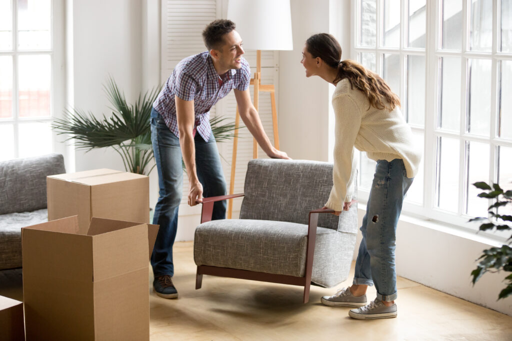 Smiling couple carrying modern chair together placing furniture moving into new home, young family discussing house improvement interior design while furnishing living room, remodeling and renovation