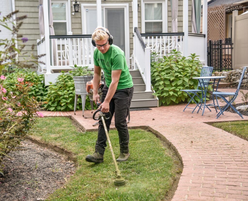 Man weed wacking a customer's grass next to a sidewalk with the house in the background.