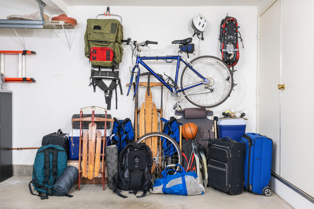 Sports and travel gear and equipment in piles in corner of messy suburban garage.