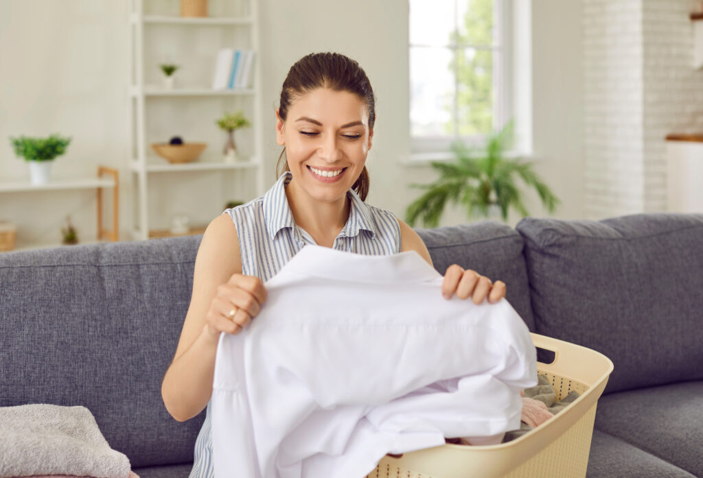 Happy housewife doing laundry. Beautiful, smiling, young woman sitting on sofa with laundry basket full of washed clothes and looking at clean, white shirt with no stains