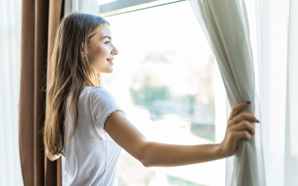 Young pretty girl opening curtains in a bedroom