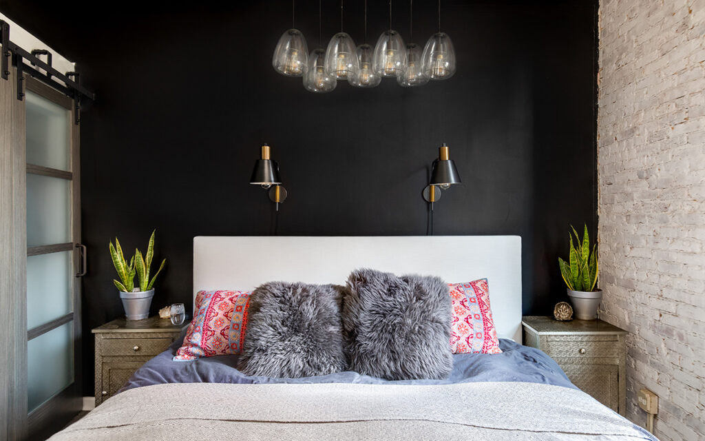 A bedroom detail with a black accent wall, black and gold lights mounted, a glass chandelier light hanging above, and colorful pillows on the bedding.