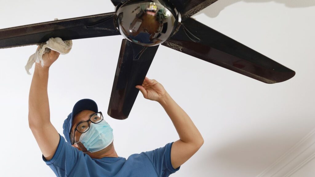 A person wearing a face mask and blue shirt is fixing a ceiling fan